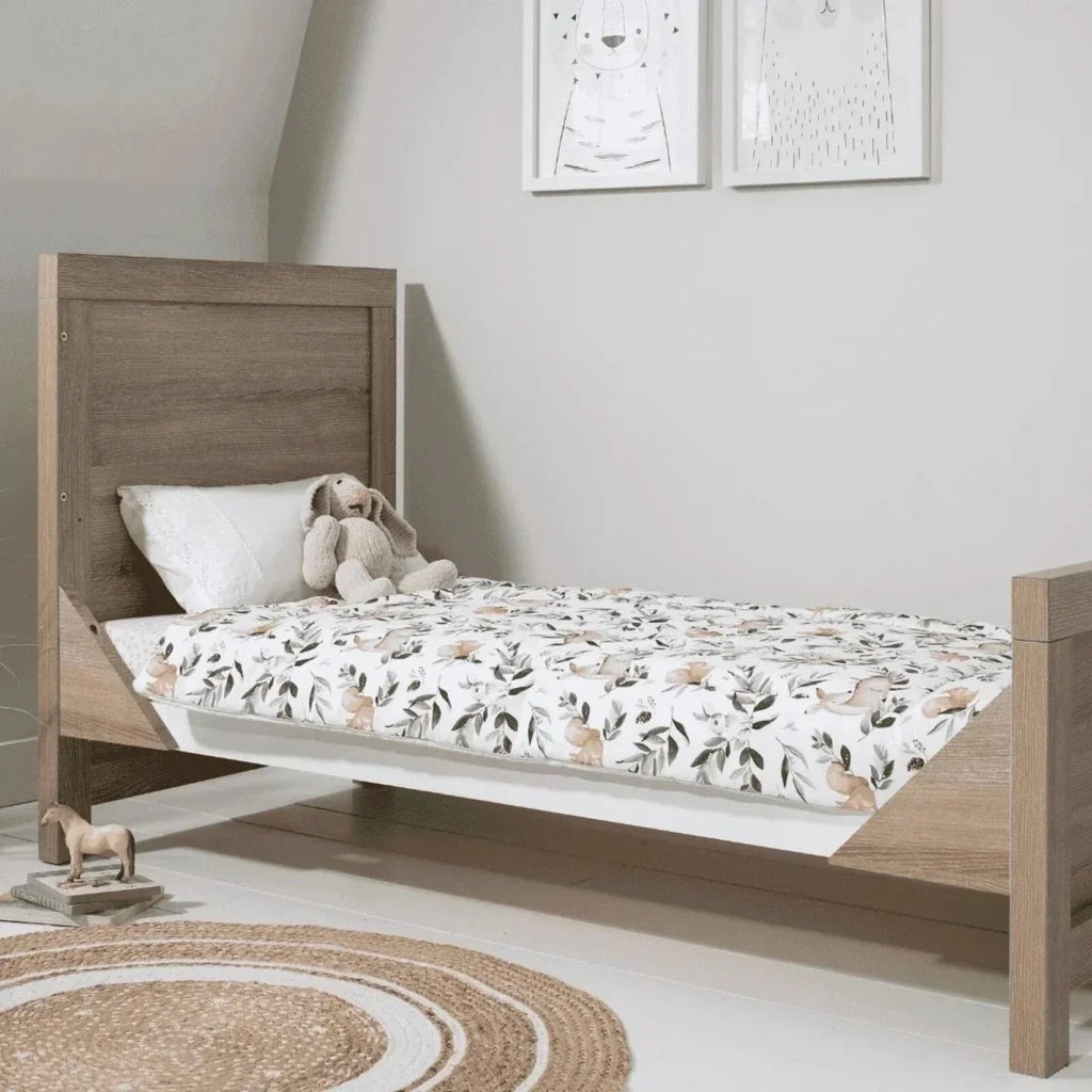 Light oak toddler bed with white patterned bedding and stuffed toys