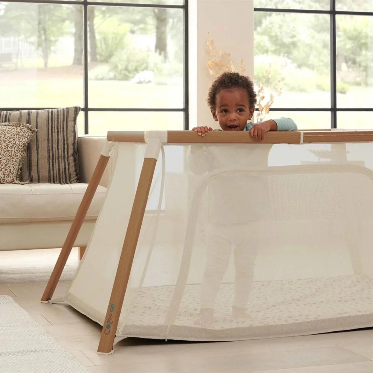 Ecru mesh playpen with light wood frame; baby peeks over the top rail.