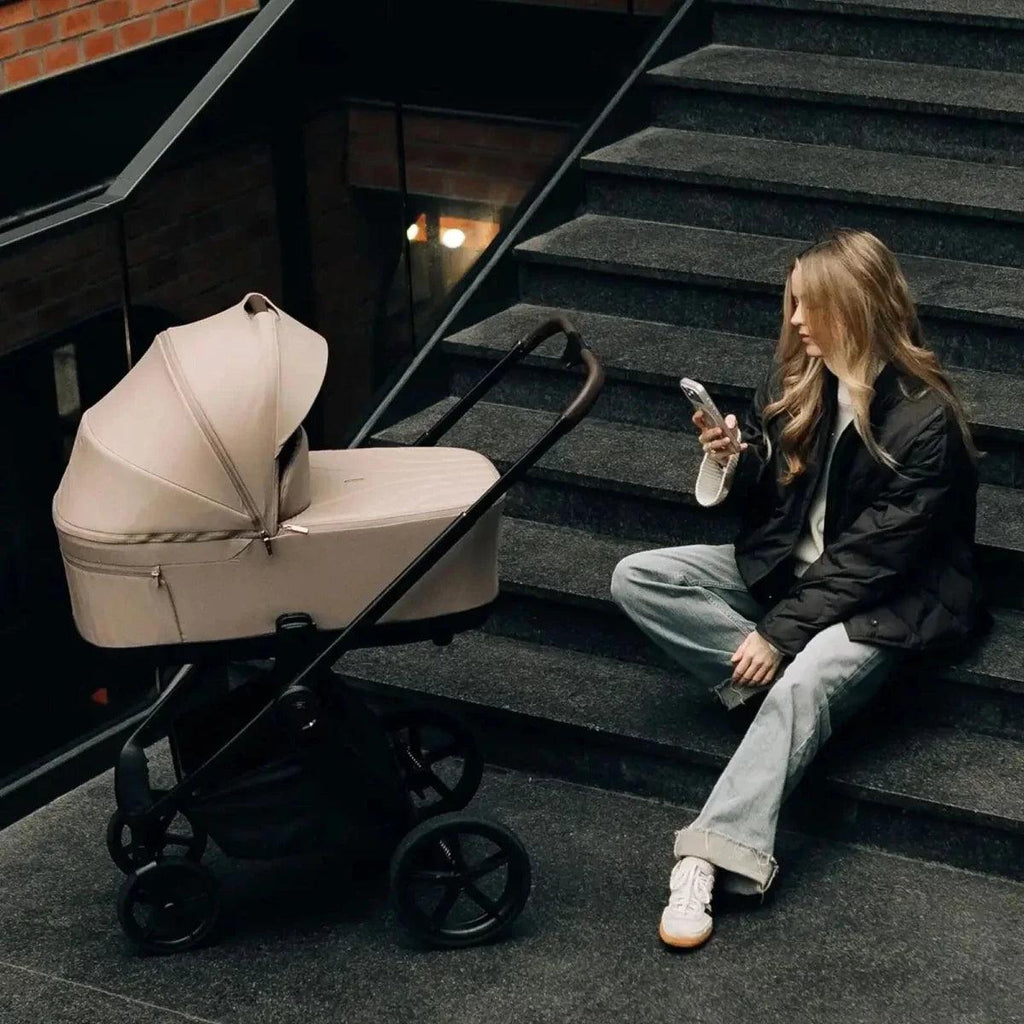 Light-colored stroller with canopy and carrycot, folded frame wheels, beside seated adult.