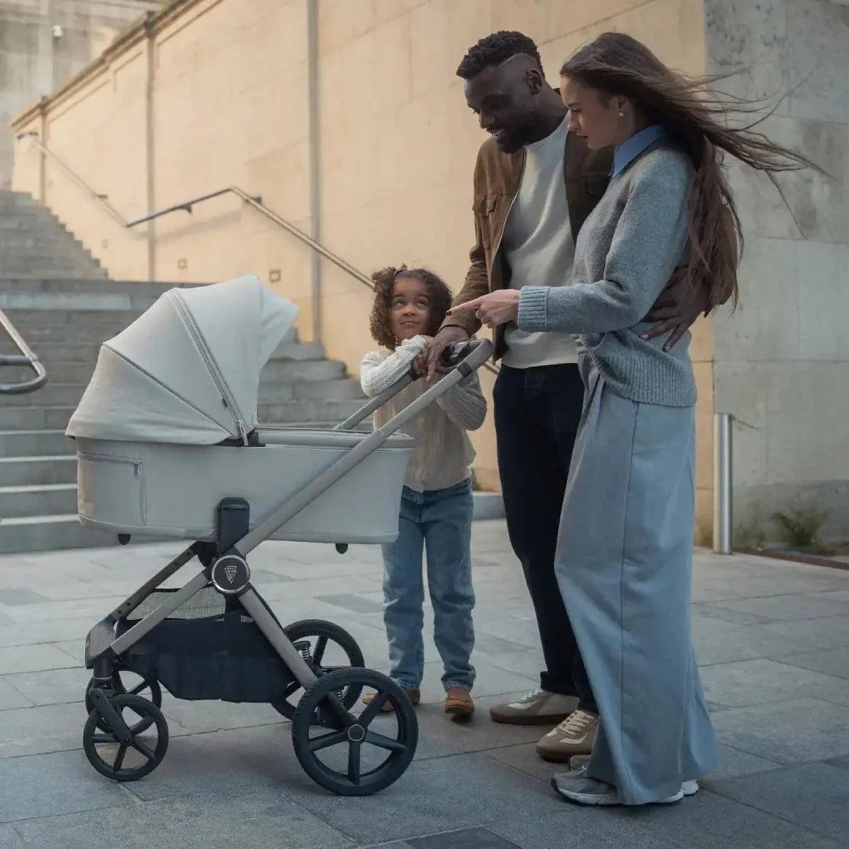 Light gray stroller carrycot with dark chassis, with family standing nearby
