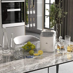Portable white ice maker on kitchen counter next to glasses, bottle and citrus fruit