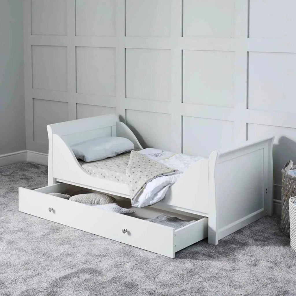 White cot bed with side rails and drawer base, shown with pillows and bedding.