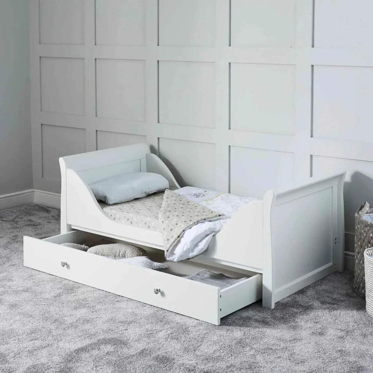 White cot bed with side rails and drawer base, shown with pillows and bedding.