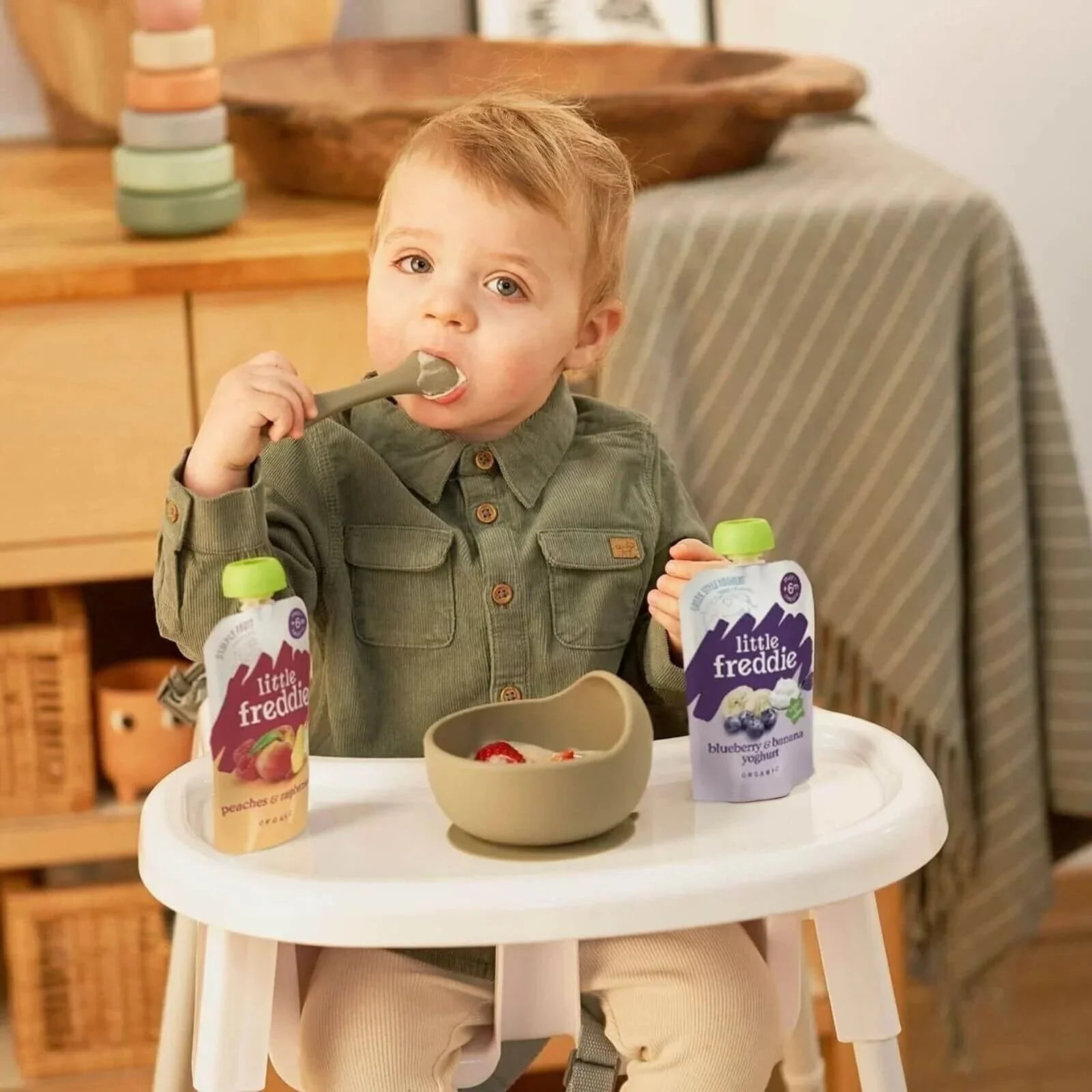 Toddler in high chair holding a Pink Lady apple yoghurt baby food pouch with green cap