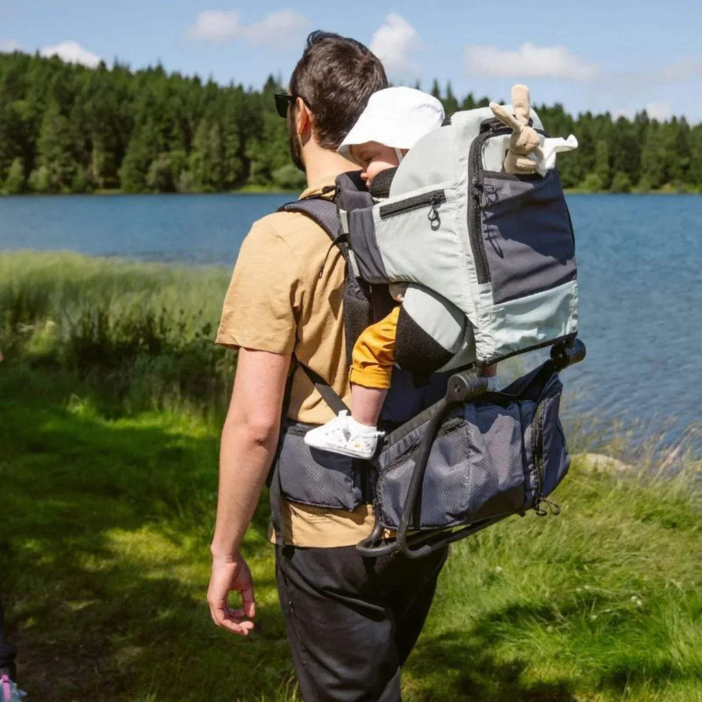 Sage green baby carrier worn by a parent, child seated with headrest and harness
