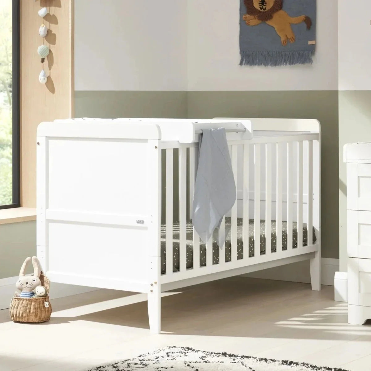 White cot bed with slatted sides and grey drape, alongside matching white dresser