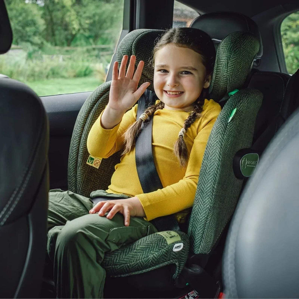 Green herringbone car seat with harness and headrest, child seated upright and waving