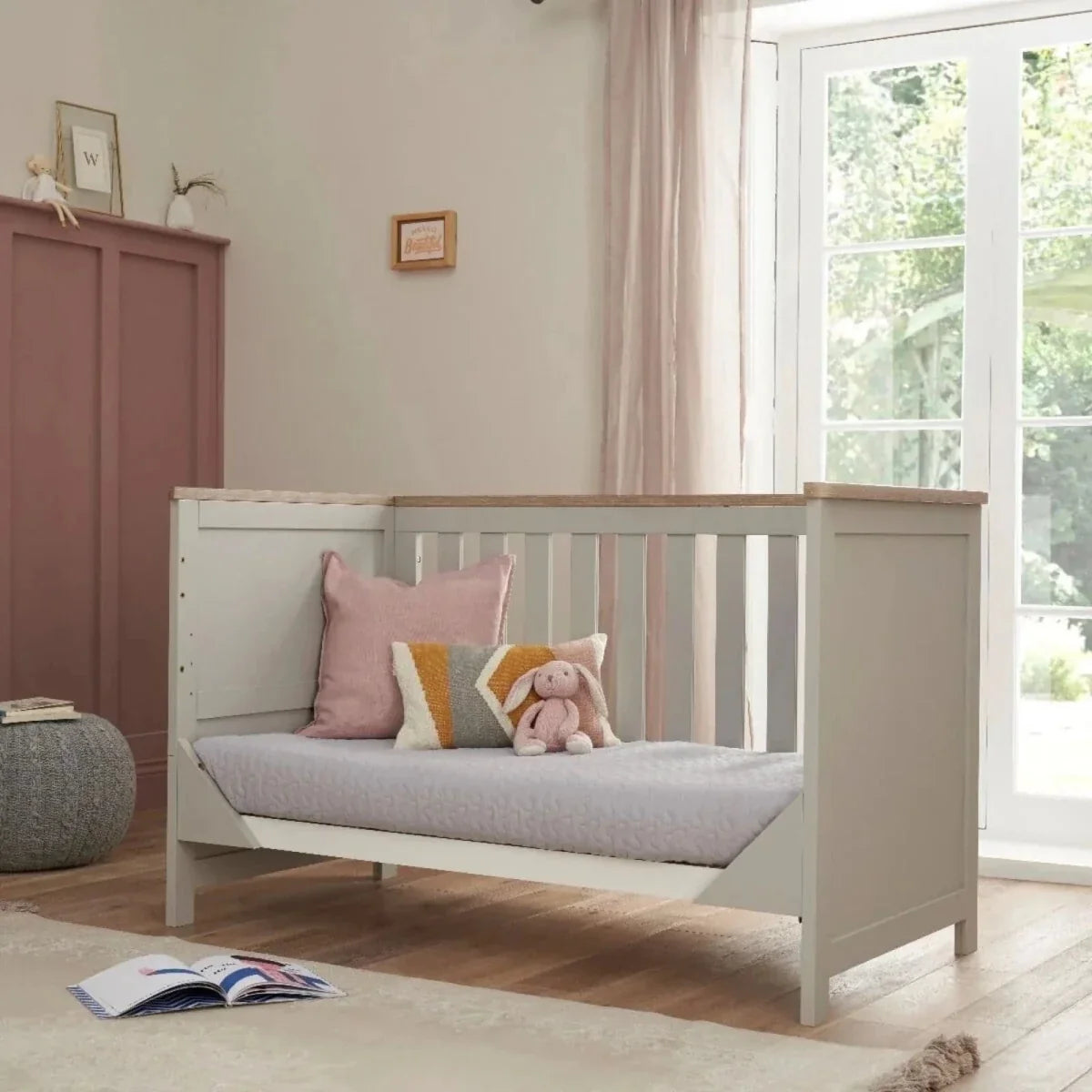 White cot bed with vertical slats and oak-toned top rail, with cushioned mattress pad