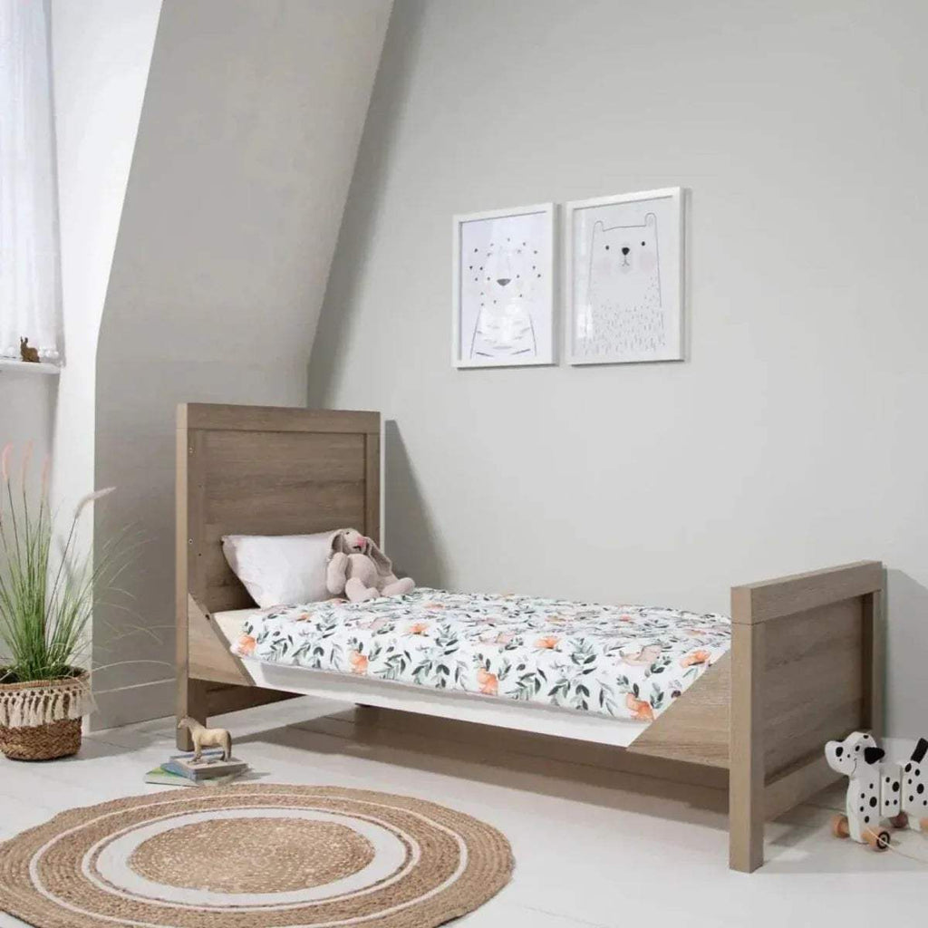 Light oak kids bed with white mattress and side rails, shown in a bedroom setting.