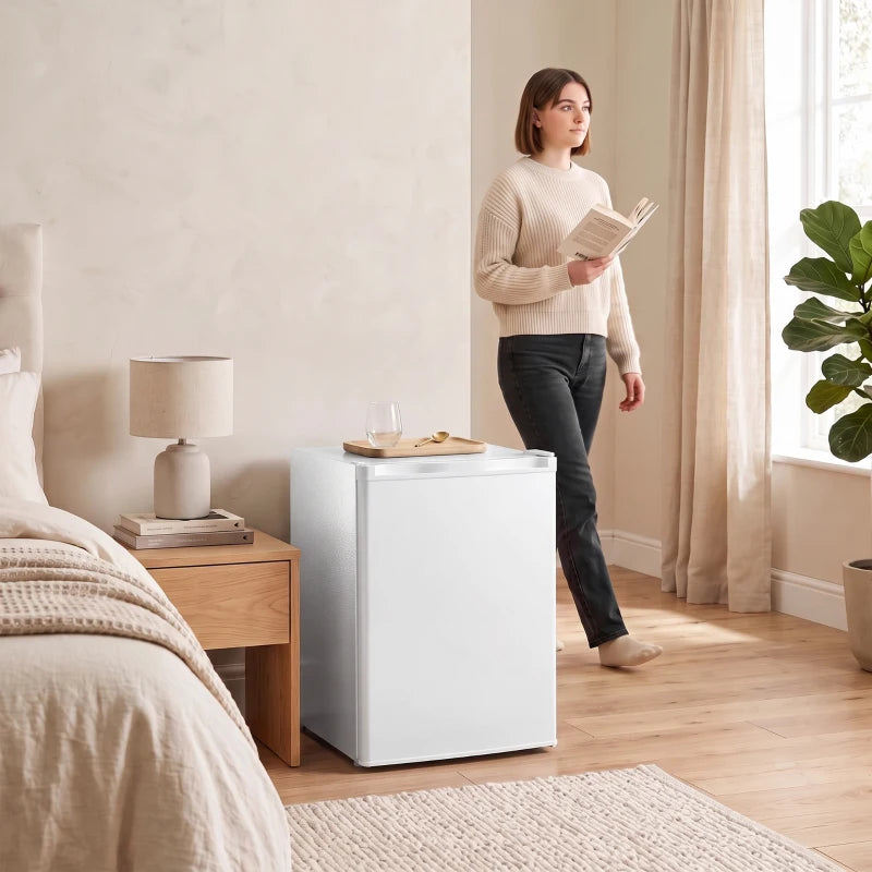 White mini fridge next to a bed in a living space, compact upright appliance