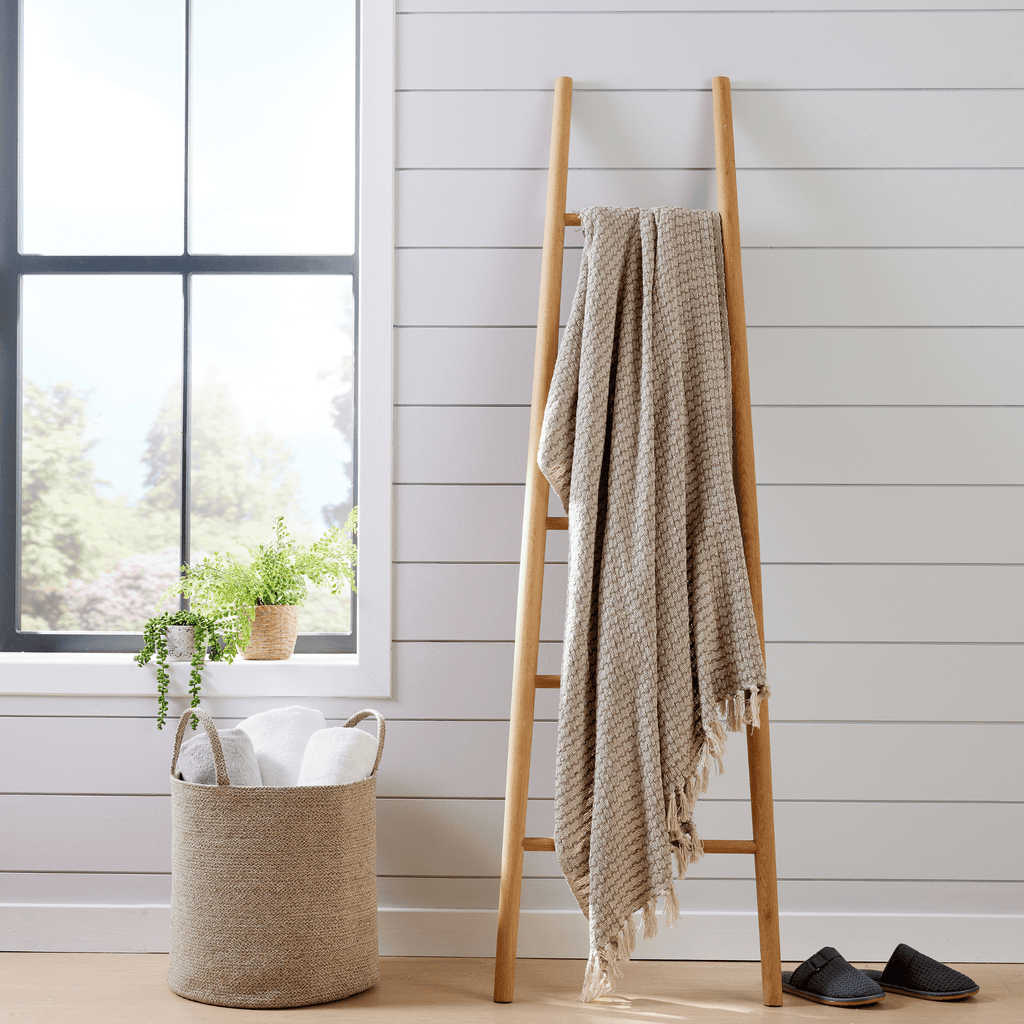 Beige woven cotton throw blanket with fringe, hanging on a wooden chair