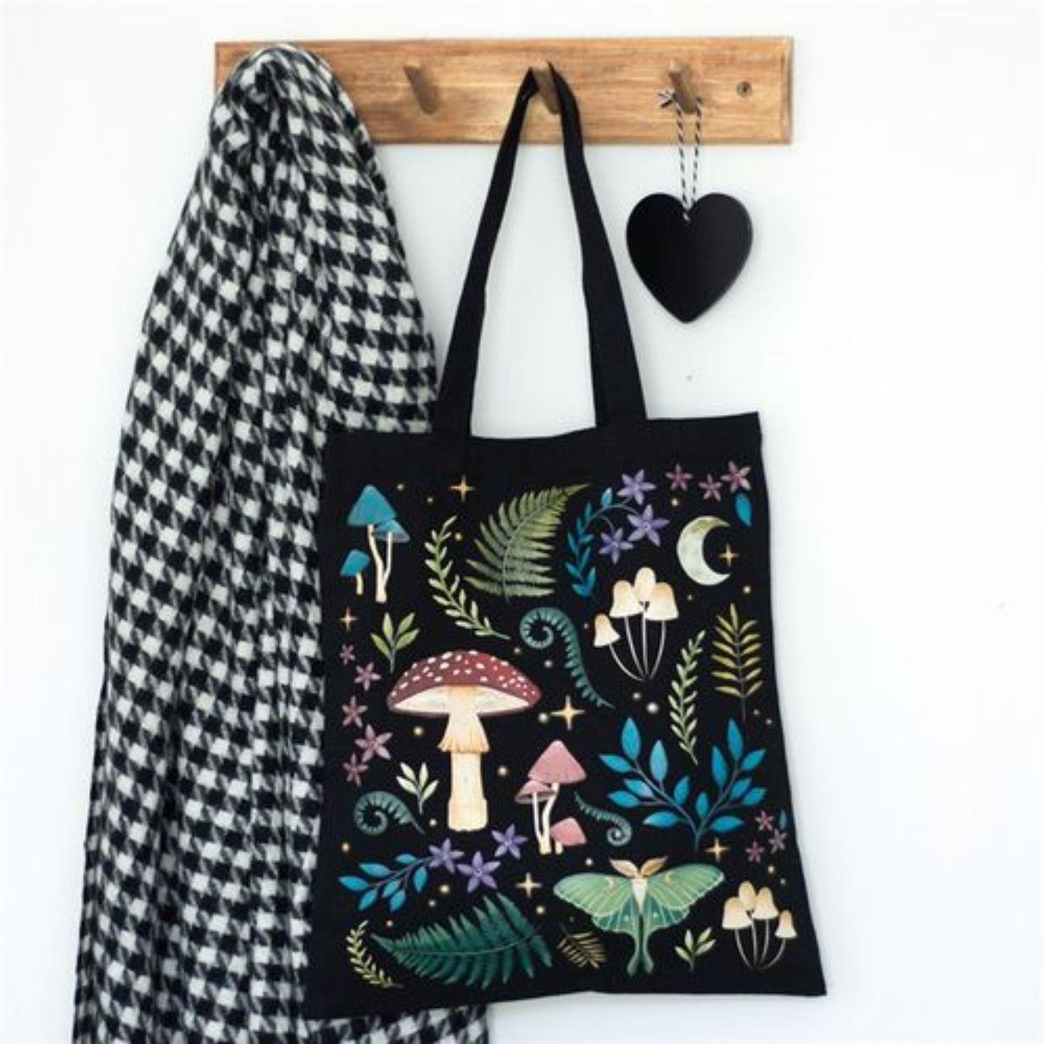 Black tote bag with nature-themed design hanging on a wooden hook with a checkered cloth and black heart.