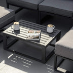 Black outdoor coffee table with slatted top beside grey cushioned ottoman seats