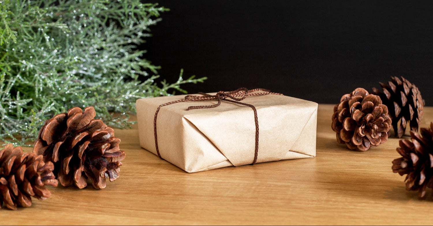 Gift wrapped in brown paper with string, surrounded by pine cones and greenery on a wooden surface.