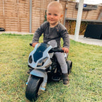 Black-and-white kids electric ride-on motorbike with foot pedal, shown outdoors with rider.